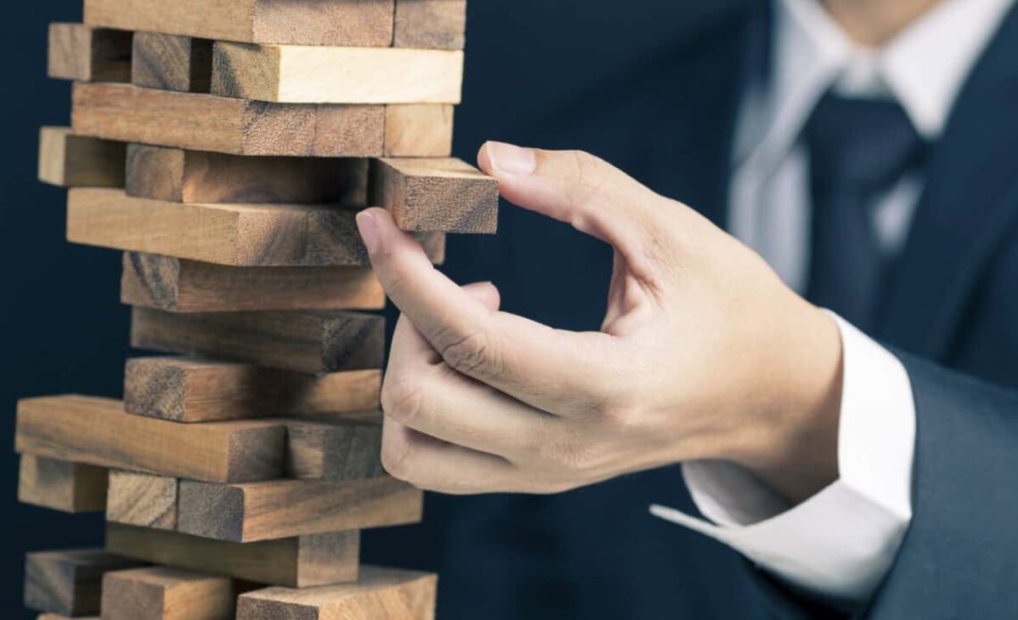 man in business suit pulls a piece out of wobbly wooden tower