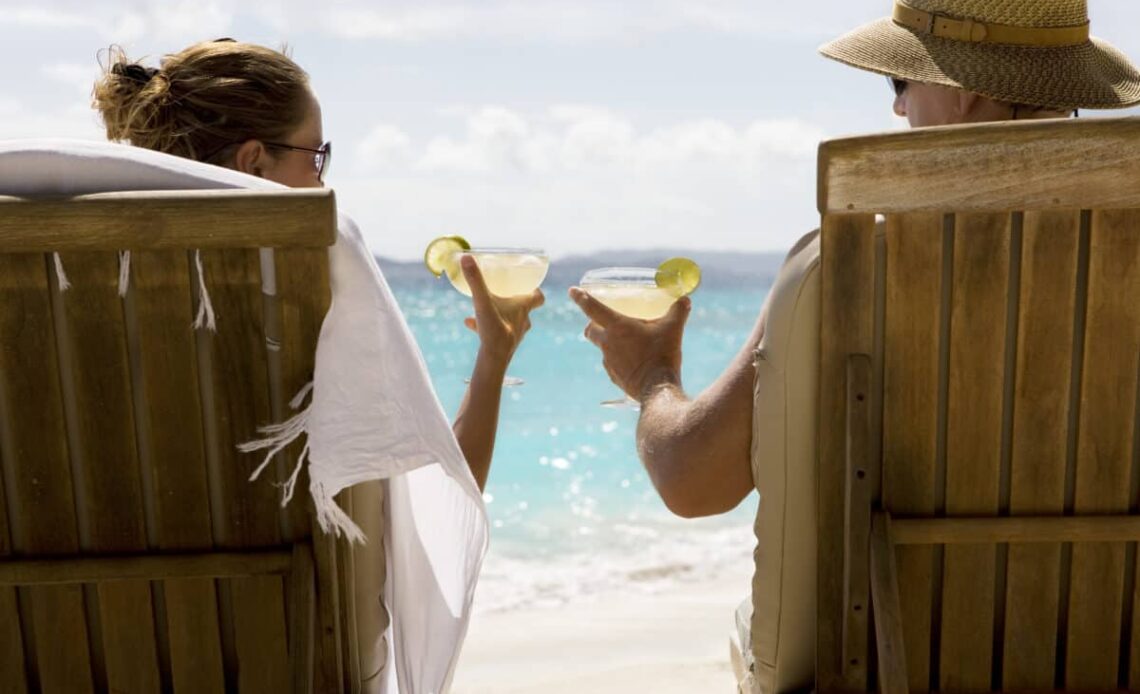 people sit in two wooden beach chairs facing the Caribbean ocean holding drinks and making a toast